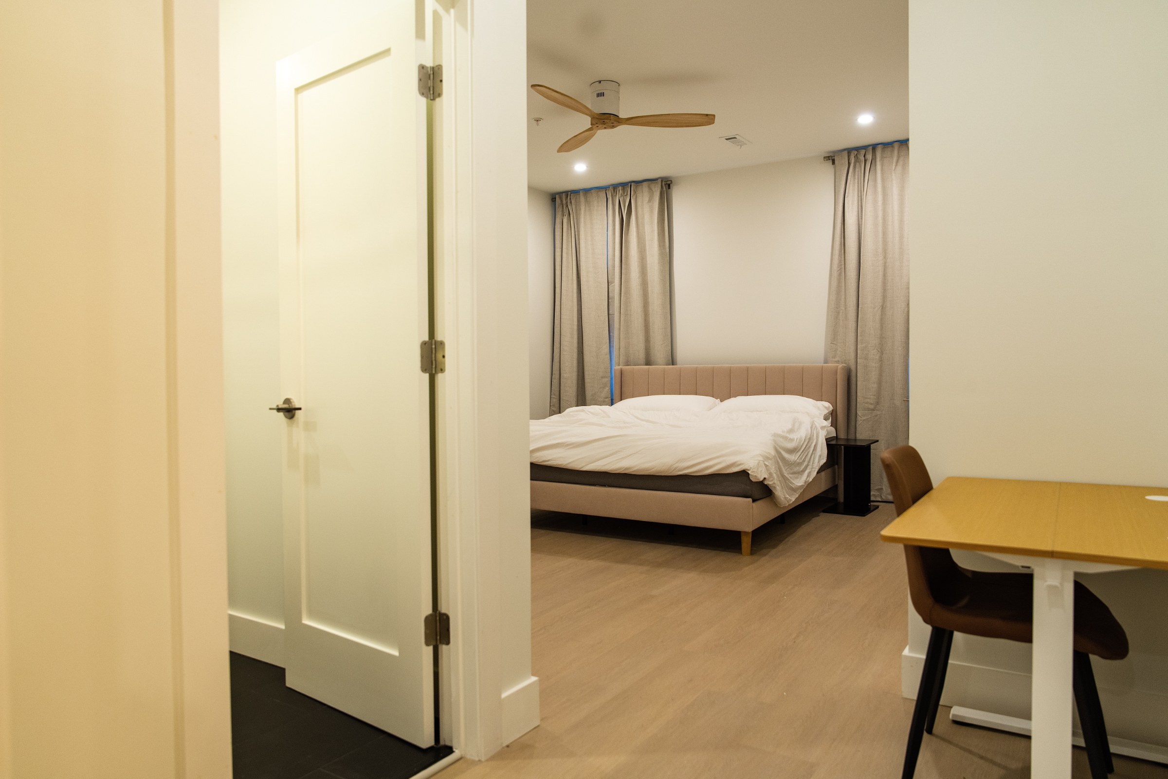 Wood-blade ceiling fan over bedroom with tan upholstered bed and gray curtains.