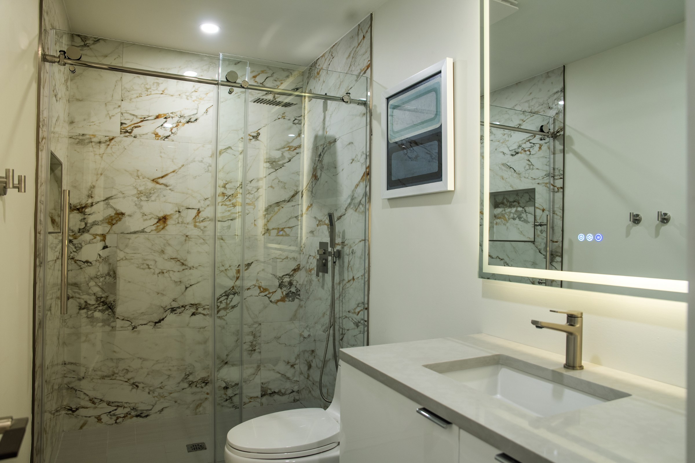 Bathroom with marble-tiled glass shower and a lit vanity mirror.